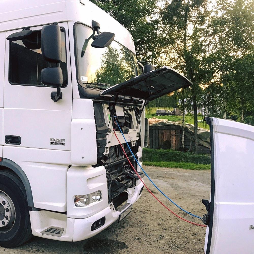 Biała ciężarówka DAF 105.460 z otwartą maską silnika, podłączona przewodami do klimatyzacji z białego busa, na tle drzew i drewnianej szopy.