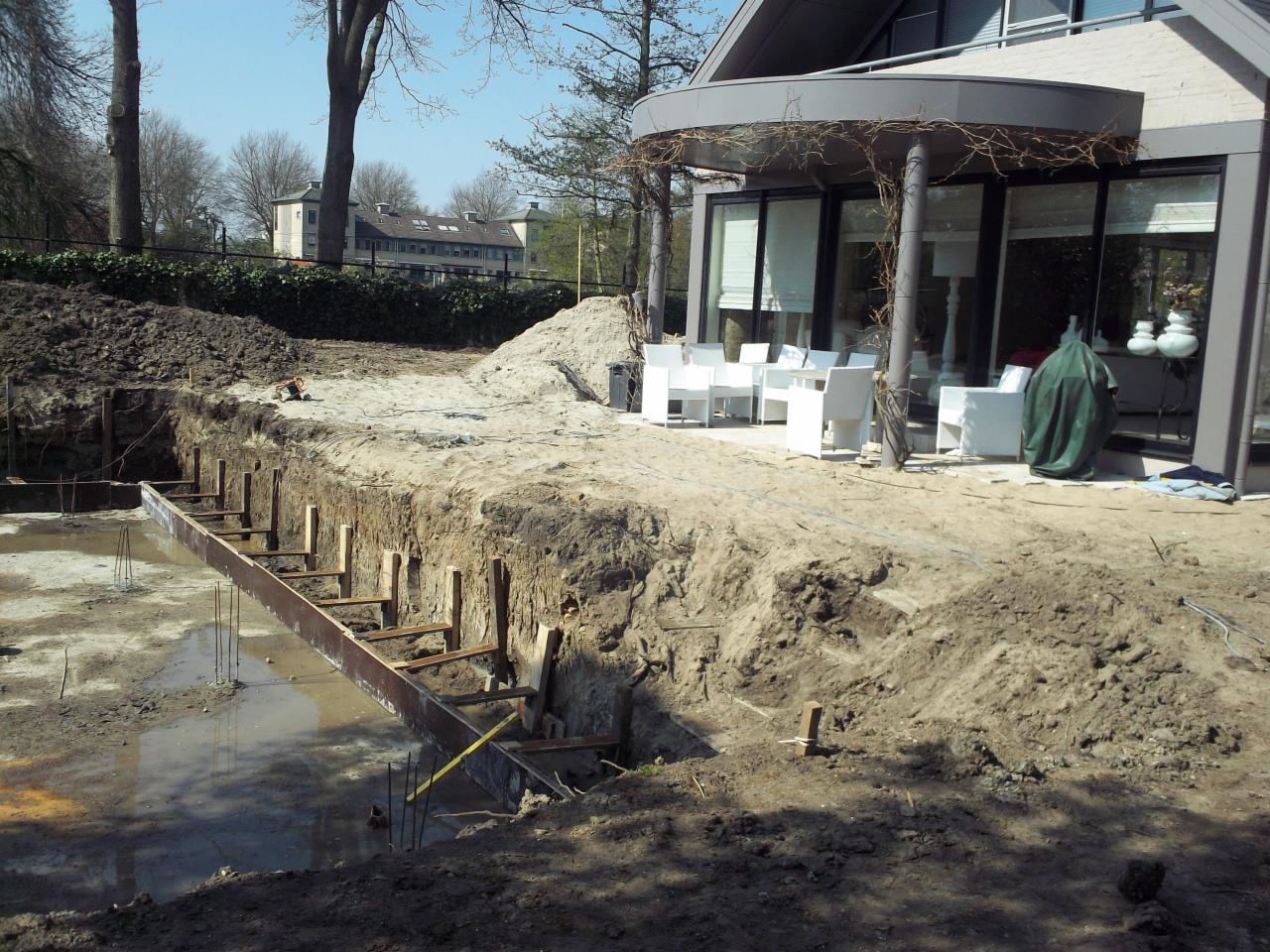 Wykop fundamentów pod budynek z widocznym szalunkiem i stojącą wodą, obok taras z białymi meblami ogrodowymi i budynek mieszkalny w tle.