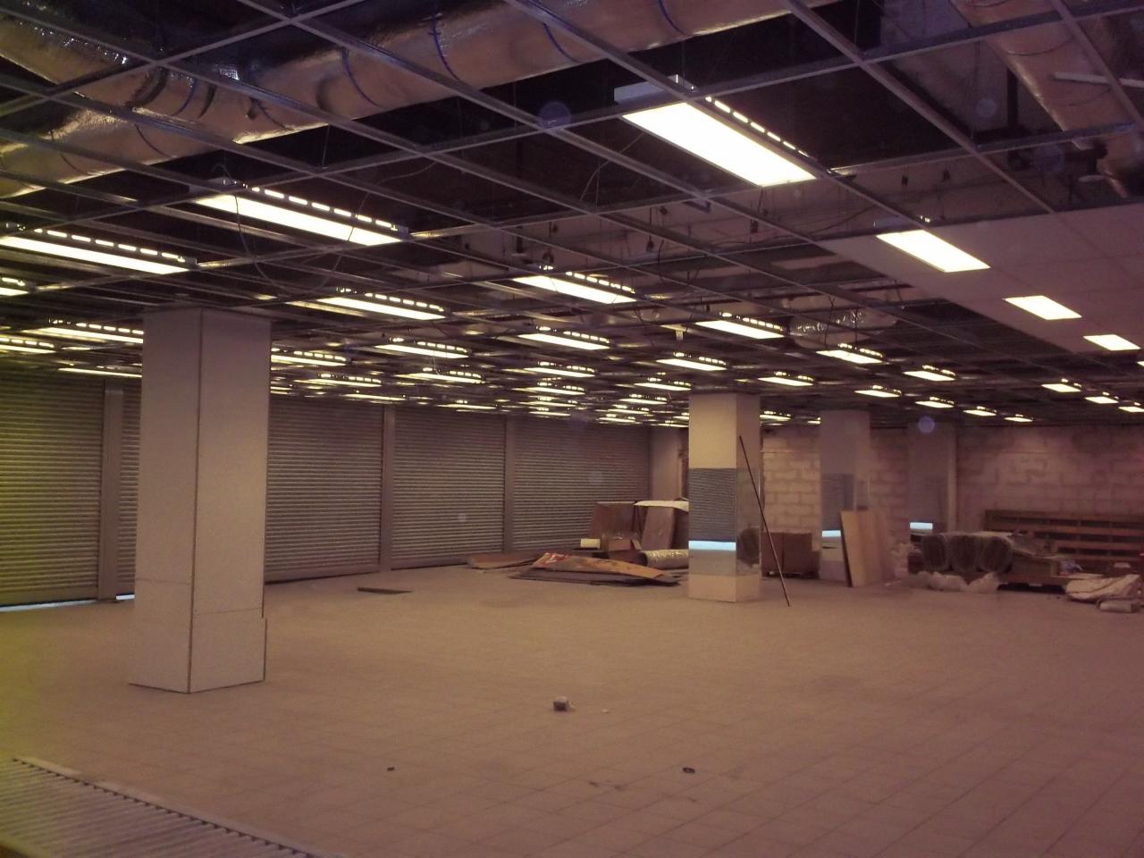 Wnętrze dużego, pustego pomieszczenia z szarymi rolowanymi drzwiami, betonowymi filarami i podwieszanym sufitem z rzędami lamp, widoczne ślady prac budowlanych.