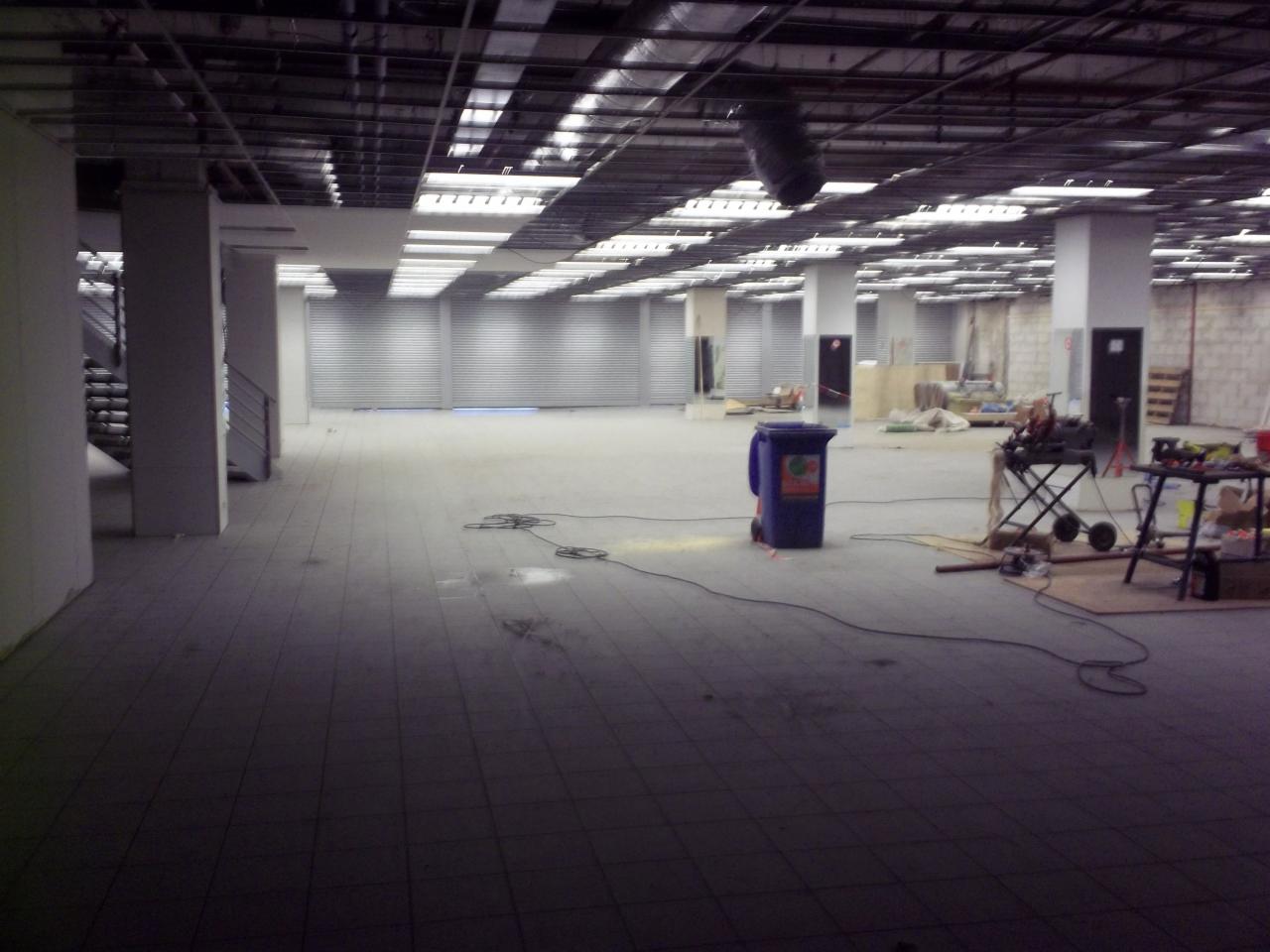 Wnętrze dużego pomieszczenia w trakcie prac wykończeniowych: widoczne kolumny, metalowe rolety w tle, niebieski kosz na śmieci, rozłożone kable i narzędzia na stole warsztatowym.