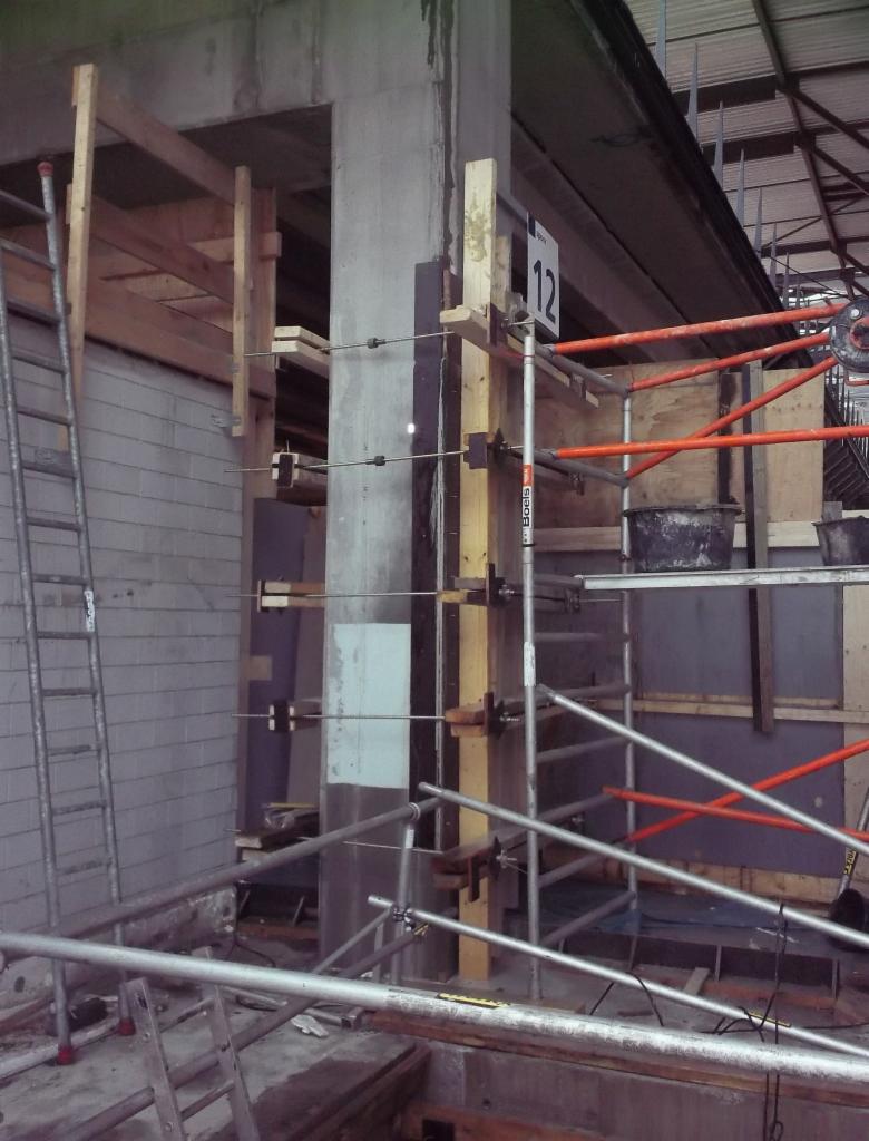 Szary betonowy filar wzmocniony drewnianymi stemplami i metalowymi ściągami w trakcie budowy, widoczne rusztowania i drabina oparta o ścianę z bloków betonowych.