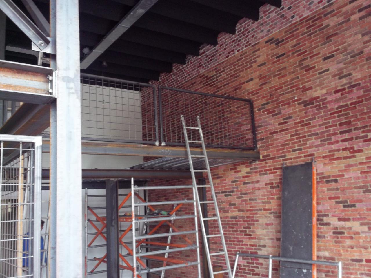 Wnętrze budynku z odsłoniętą czerwoną cegłą, metalowymi konstrukcjami, schodami i rusztowaniem, ukazujące etap prac budowlanych.