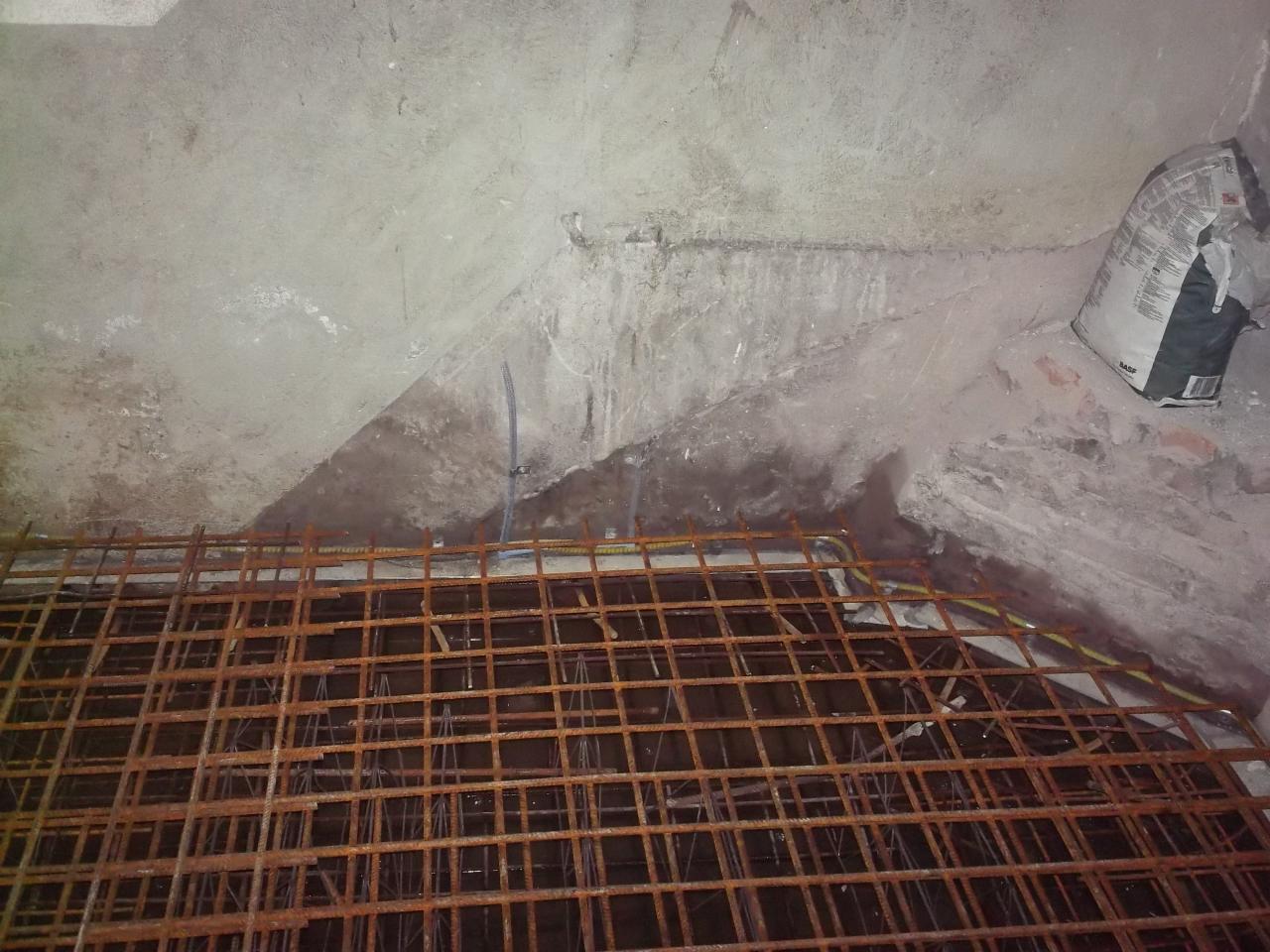 Przygotowanie zbrojenia pod wylewkę betonową w pomieszczeniu z surowymi ścianami, widoczny worek z zaprawą budowlaną w rogu.