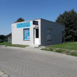 Narożny budynek biura kredytowo-ubezpieczeniowego CUK z widocznym szyldem informacyjnym i plakatem w oknie, otoczony zielenią i brukowaną drogą.