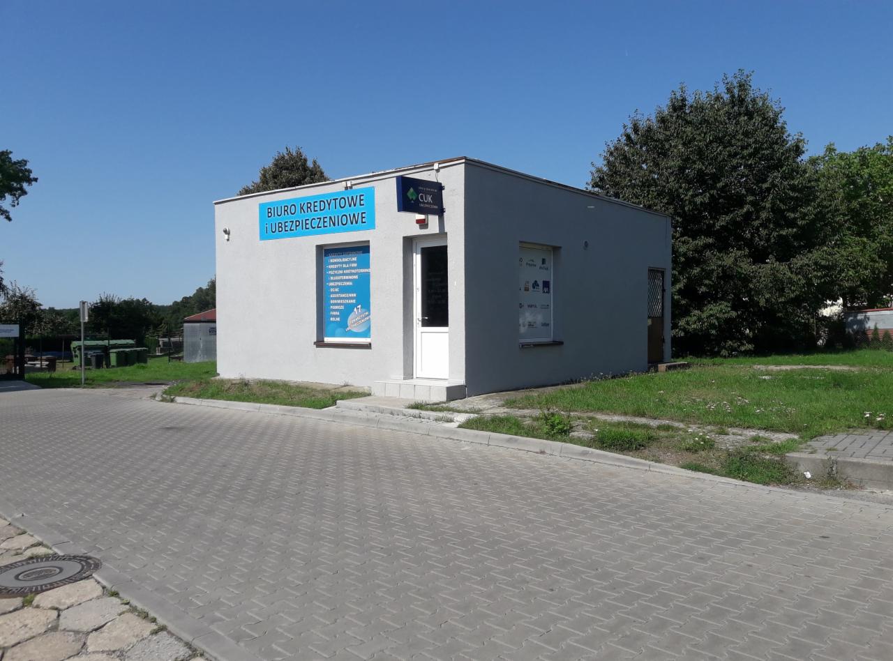 Narożny budynek biura kredytowo-ubezpieczeniowego CUK z widocznym szyldem informacyjnym i plakatem w oknie, otoczony zielenią i brukowaną drogą.