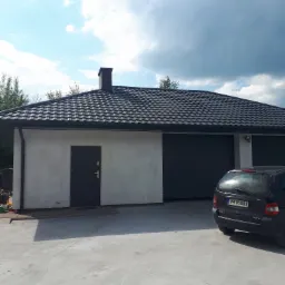 Nowoczesny budynek garażowy z ciemną, metalową dachówką falistą, czarnymi rynnami i obróbkami blacharskimi, widok z zewnątrz w słoneczny dzień.