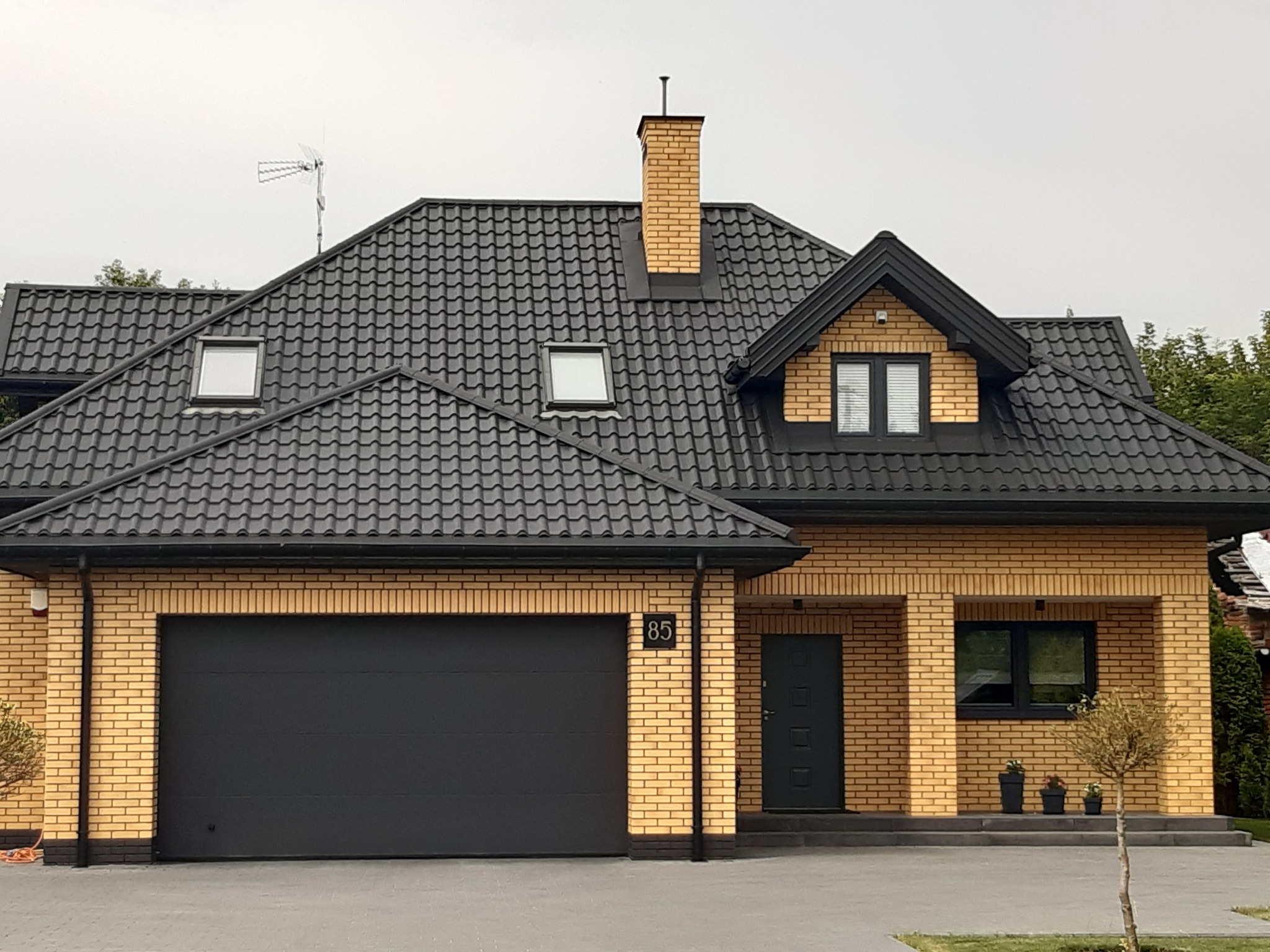 Dom jednorodzinny z czarną dachówką ceramiczną karpiówką, żółtą cegłą klinkierową, czarną bramą garażową i numerem domu 85.
