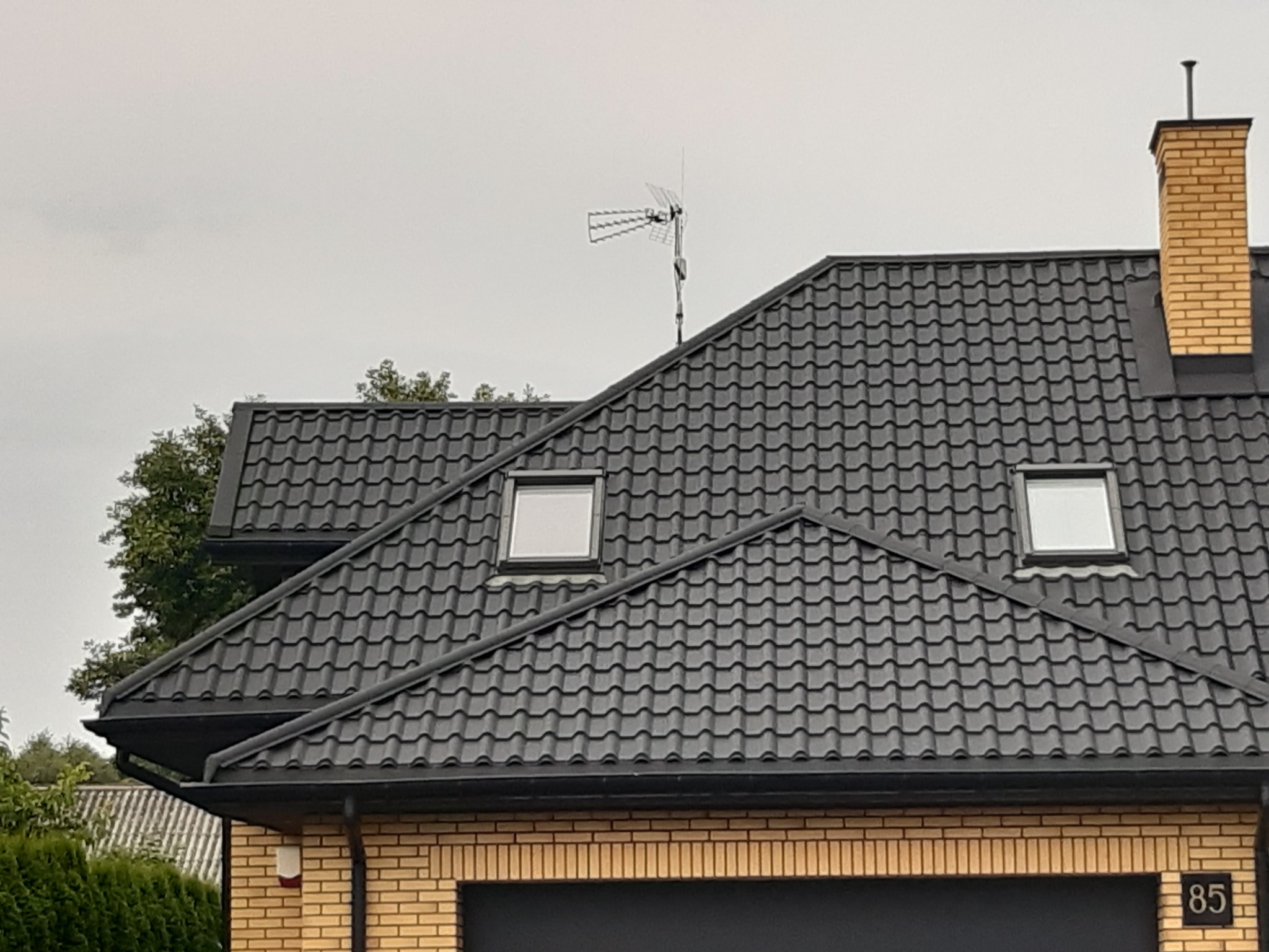 Czarny dach z dachówki ceramicznej z dwoma oknami dachowymi, anteną telewizyjną i kominem z żółtej cegły na tle pochmurnego nieba.