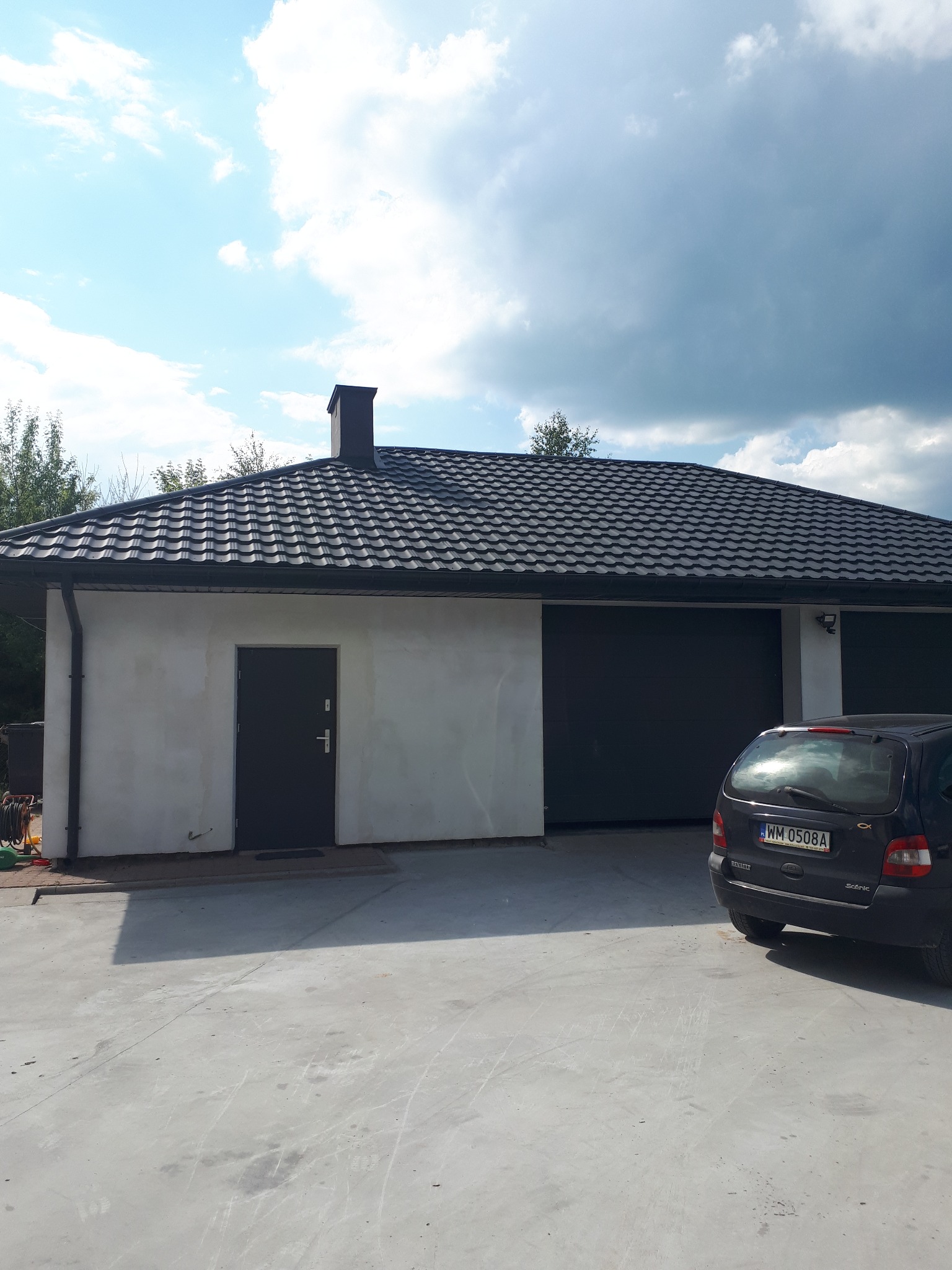 Nowoczesny budynek garażowy z ciemną, metalową dachówką falistą, czarnymi rynnami i obróbkami blacharskimi, widok z zewnątrz w słoneczny dzień.