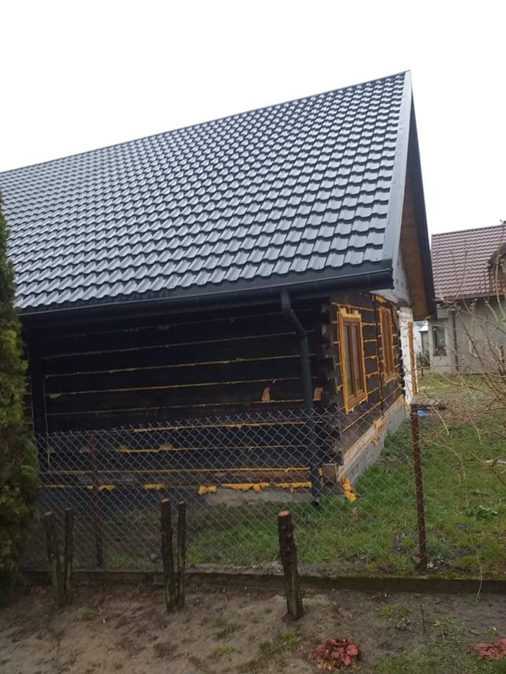Dom z bali z nowym, grafitowym dachem z blachy trapezowej i rynnami, widoczne okna w trakcie montażu i siatka ogrodzeniowa.