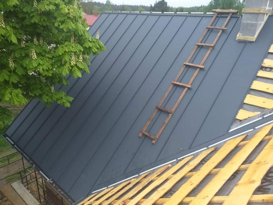 Nowy, grafitowy dach z blachy na drewnianej konstrukcji, z drewnianą drabiną opartą o połać, widok z góry, obok kwitnące drzewo.