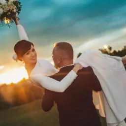 Pan młody niesie uśmiechniętą pannę młodą na rękach, trzymającą bukiet ślubny w górze, w promieniach zachodzącego słońca na tle zieleni.