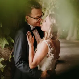 Para młoda w objęciach, panna młoda w sukni z koronkowymi aplikacjami na plecach, pan młody w czarnym smokingu i okularach, romantyczny portret w zielonej scenerii.