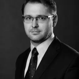 Portret biznesowy mężczyzny w okularach i garniturze, czarno-biały, z subtelnym uśmiechem i wzorem na krawacie.