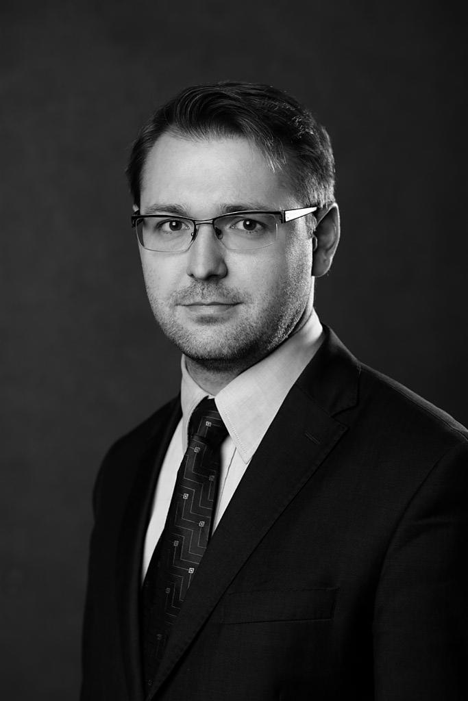 Portret biznesowy mężczyzny w okularach i garniturze, czarno-biały, z subtelnym uśmiechem i wzorem na krawacie.