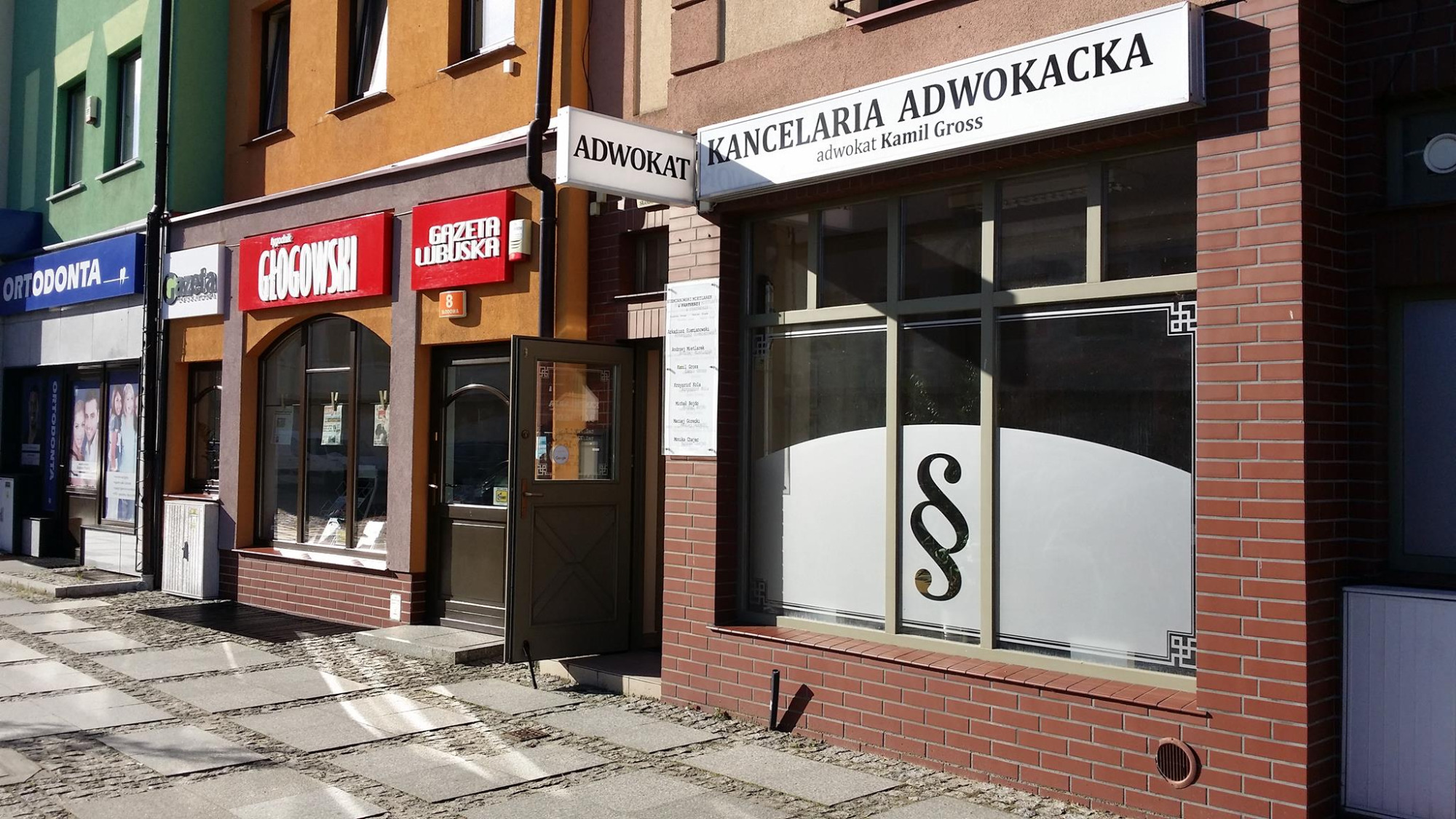 Fasada budynku z szyldem 'Kancelaria Adwokacka adwokat Kamil Gross' i znakiem paragrafu na oknie, widok z zewnątrz na ulicę z chodnikiem wyłożonym kostką.