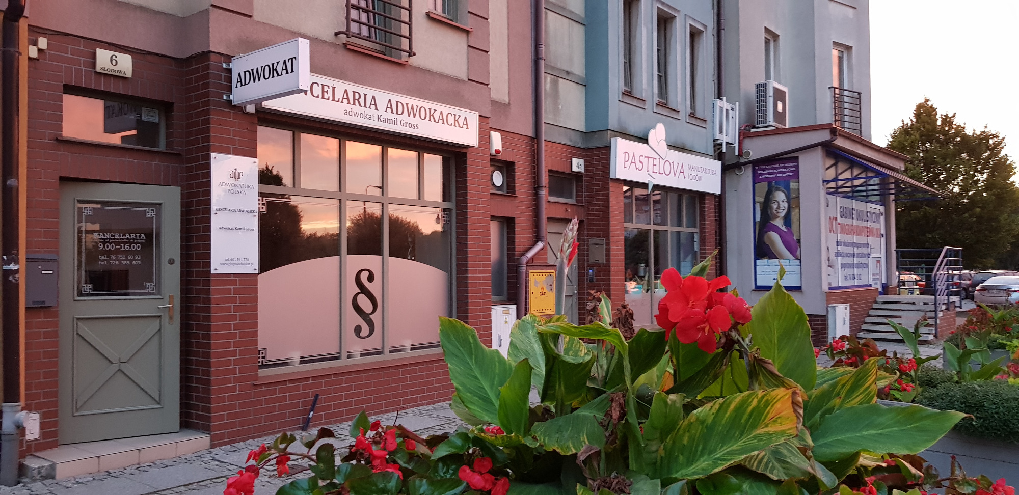 Fasada budynku z szyldami: Adwokat, Celaria Adwokacka adwokat Kamil Gross, oraz tabliczką z godzinami otwarcia kancelarii. W oknie widoczny znak paragrafu. Na pierwszym planie kwiaty.