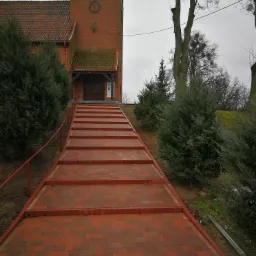 Czerwona kostka brukowa ułożona w schody prowadzące do ceglanego kościoła z wieżą zegarową, otoczonego zielenią.