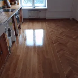 Ukończona podłoga w kuchni z jasnego parkietu ułożonego w jodełkę na części powierzchni i w proste deski na pozostałej części, z widocznym fragmentem kuchennej zabudowy i kaloryferem przy oknie.