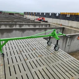 Zielony wózek transportowy na betonowych płytach z wyciętymi szczelinami, w tle widoczne ściany fundamentowe i maszyna budowlana, pochmurne niebo.