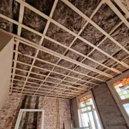 Drewniana konstrukcja sufitu z izolacją z wełny mineralnej, widoczne fragmenty ceglanej ściany i okno ze szprosami i witrażem w trakcie remontu.