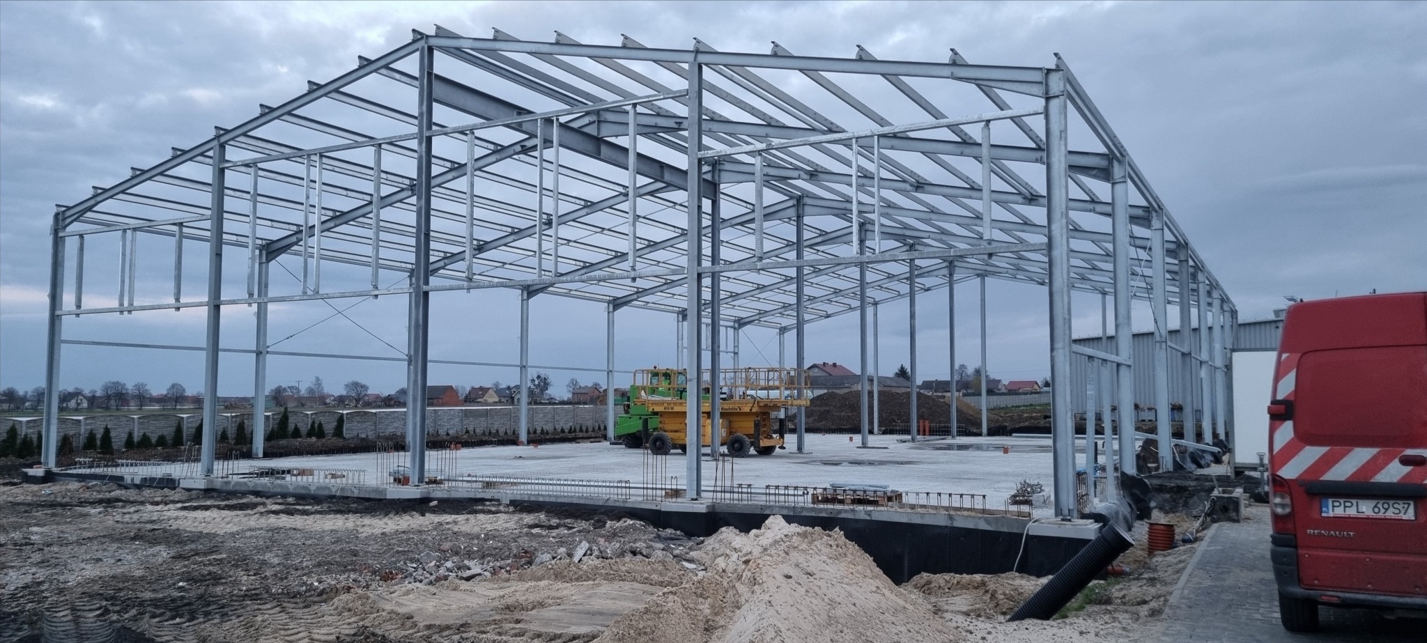 Stalowa konstrukcja hali w trakcie montażu, widoczny fundament i podłoże przygotowane pod wylewkę, na placu budowy żółty podnośnik, w tle zabudowa jednorodzinna.