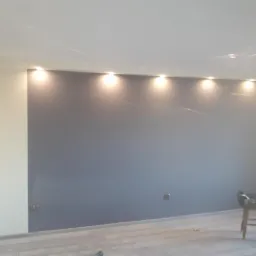 Świeżo pomalowana szara ściana z oświetleniem punktowym w pomieszczeniu w Książu Wielkopolskim, widoczne drewniane panele podłogowe i krzesło z narzędziami.