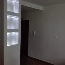 Nowoczesna ściana działowa z wnękami oświetlonymi LED, ciemna podłoga, białe ściany i drzwi w tle.