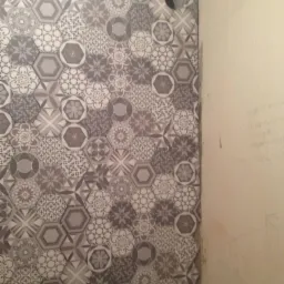 Ściana w rogu pomieszczenia wyłożona dekoracyjnymi, heksagonalnymi płytkami w odcieniach szarości i bieli; widoczny otwór na instalację.