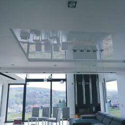 WilczykBud - Nowoczesny salon z błyszczącym, lustrzanym sufitem podwieszanym, który odbija meble i krajobraz za oknem; widoczne duże okna z widokiem na wzgórza i zabudowania.