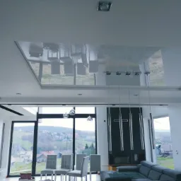 Nowoczesny salon z błyszczącym, lustrzanym sufitem podwieszanym, który odbija meble i krajobraz za oknem; widoczne duże okna z widokiem na wzgórza i zabudowania.