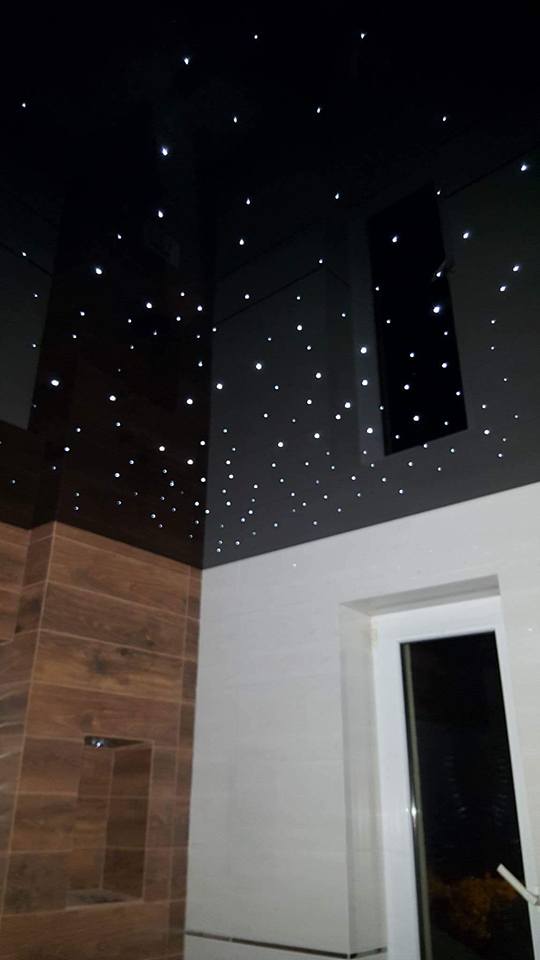 Nowoczesna łazienka z podświetlanym sufitem imitującym gwiazdy, brązowe płytki imitujące drewno i białe płytki ścienne, widoczne okno.