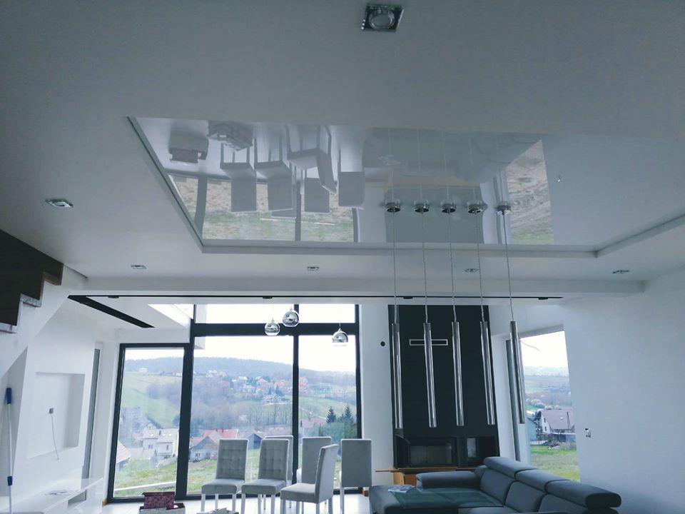 Nowoczesny salon z błyszczącym, lustrzanym sufitem podwieszanym, który odbija meble i krajobraz za oknem; widoczne duże okna z widokiem na wzgórza i zabudowania.