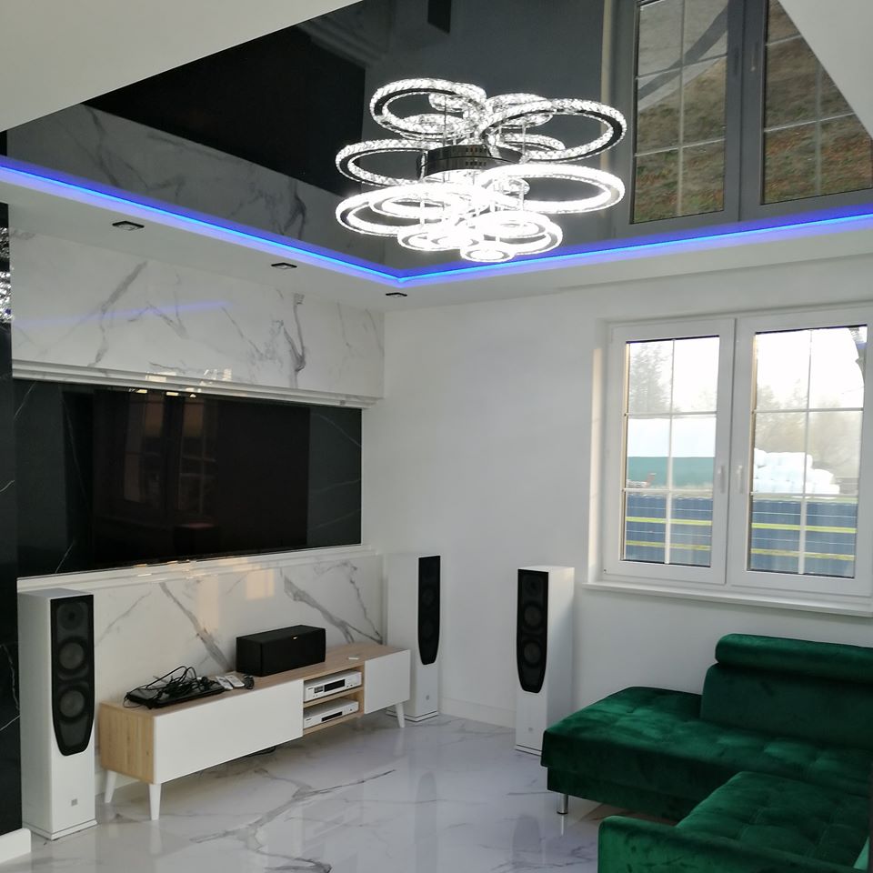 Salon z czarnym, lustrzanym sufitem podwieszanym, oświetleniem LED i nowoczesnym żyrandolem, marmurowe wykończenie ściany za telewizorem, białe głośniki podłogowe i zielona sofa narożna.