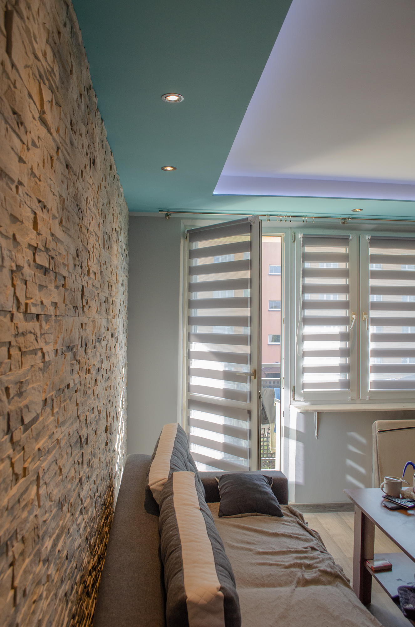 Salon z dekoracyjną ścianą z kamienia, roletami dzień-noc w oknach balkonowych i podwieszanym sufitem z oświetleniem punktowym oraz ledowym.