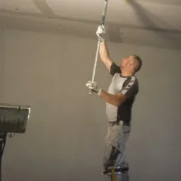 Mężczyzna na szczudłach szpachluje sufit za pomocą narzędzia z długim uchwytem, widoczna szpachlowana powierzchnia sufitu i wiadro ze szpachlą.