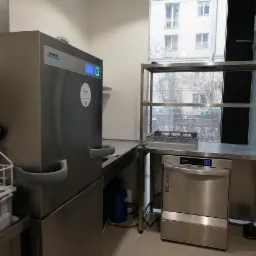 Profesjonalna zmywarka gastronomiczna Winterhalter UC Series w jasnym pomieszczeniu z oknem, widoczna kratka z naczyniami i niebieski kanister obok większej zmywarki Winterhalter.