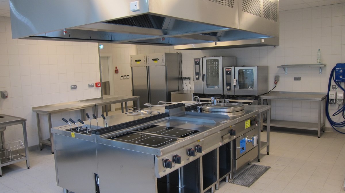 Profesjonalna kuchnia gastronomiczna ze stali nierdzewnej, widoczne piece konwekcyjne, frytownica, płyty grzewcze, okap i blaty robocze, białe kafelki na ścianach.