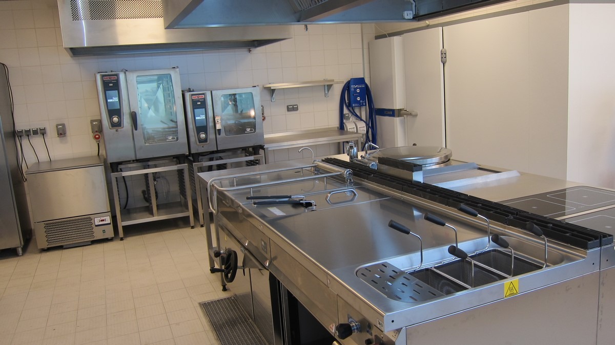Profesjonalna kuchnia gastronomiczna ze stali nierdzewnej, widoczne piece konwekcyjno-parowe, stanowisko do smażenia frytek z czterema koszami i płyta grzewcza, całość w jasnym pomieszczeniu...