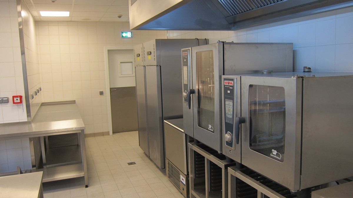 Wnętrze profesjonalnej kuchni gastronomicznej z rzędem pieców konwekcyjno-parowych marki Rational, stalowymi stołami roboczymi i chłodziarkami, oświetlone jasnym, górnym światłem.