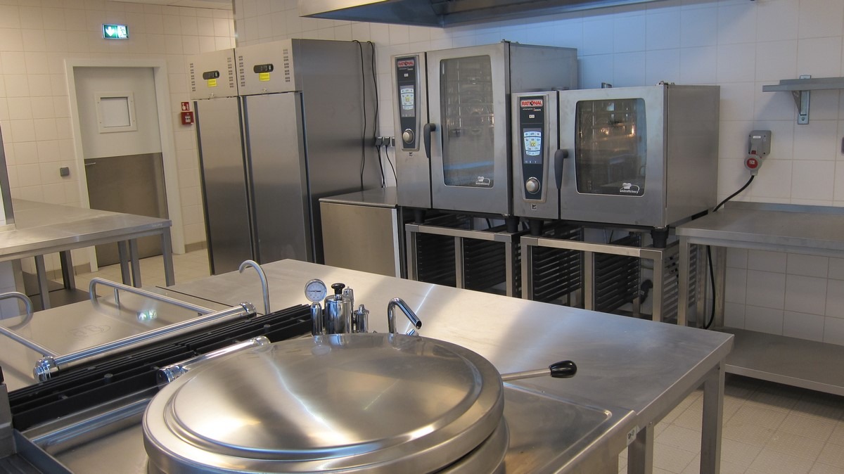 Profesjonalna kuchnia gastronomiczna ze stali nierdzewnej, widoczne piece konwekcyjno-parowe, chłodziarki i stół roboczy z dużym garnkiem ciśnieniowym z manometrem, oświetlenie sufitowe, płytki...