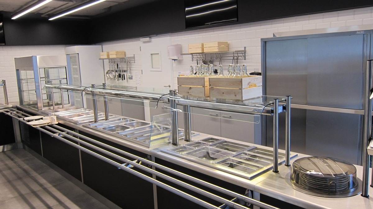 Profesjonalne wyposażenie bufetu w restauracji: stalowe pojemniki GN w różnych rozmiarach, szklane osłony, stos talerzy pod przykryciem, drewniane skrzynki z butelkami na tle białych płytek.