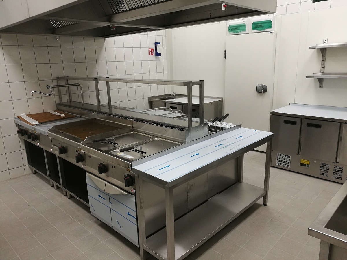 Profesjonalna kuchnia gastronomiczna ze stali nierdzewnej, widoczny grill, frytownica i stół roboczy, w tle białe kafelki i drzwi chłodnicze.