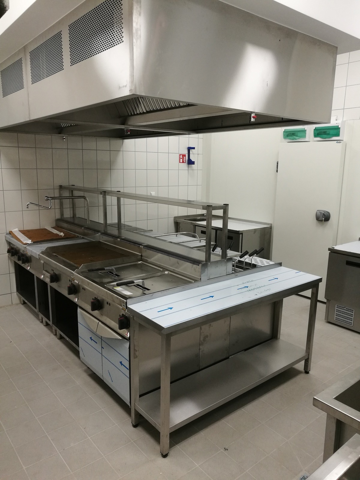 Profesjonalna kuchnia gastronomiczna ze stali nierdzewnej, widoczny piec, frytownica i stół roboczy pod okapem, płytki na ścianach.