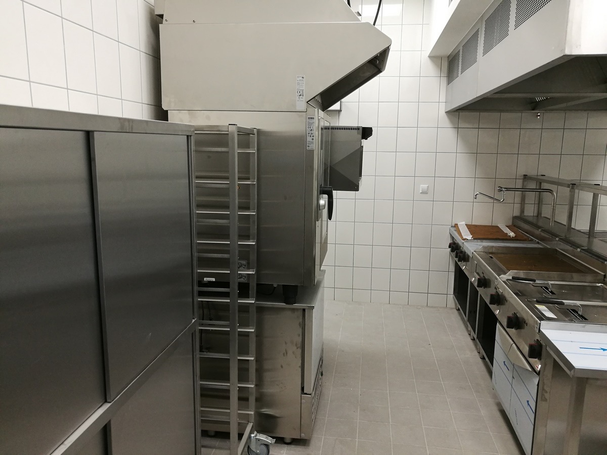 Profesjonalna kuchnia gastronomiczna ze stali nierdzewnej: piec konwekcyjno-parowy z wózkiem transportowym, frytownica i szafki w pomieszczeniu wyłożonym białymi kafelkami.