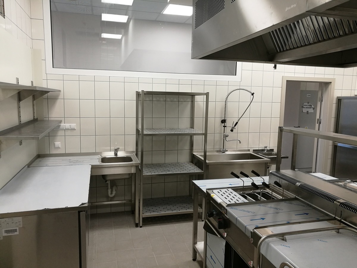 Profesjonalna kuchnia gastronomiczna z nierdzewnymi urządzeniami, w tym frytownicą, zlewem z elastyczną wylewką i metalowym regałem magazynowym, wyłożona jasnymi płytkami.