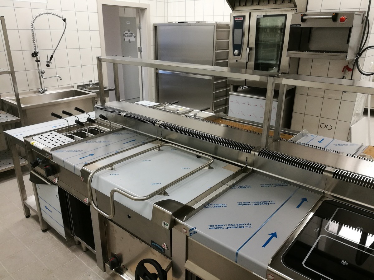 Profesjonalna kuchnia ze stali nierdzewnej, wyposażona w frytownice, piec konwekcyjny i stanowisko do mycia naczyń, całość w jasnym pomieszczeniu z białymi kafelkami.