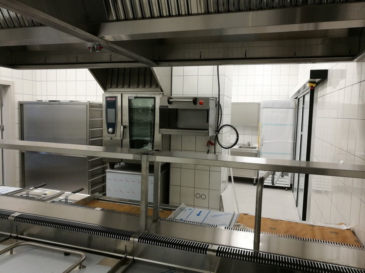 Profesjonalna kuchnia gastronomiczna z piecem konwekcyjno-parowym Rational, grillem, chłodziarką i elementami ze stali nierdzewnej, widok z przodu, białe płytki na ścianach.