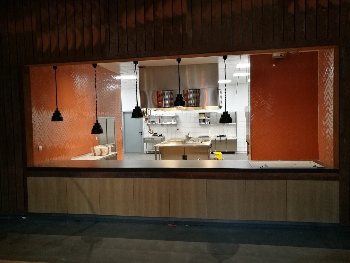 Widok na profesjonalną kuchnię gastronomiczną z pomarańczowymi ścianami w jodełkę i wiszącymi czarnymi lampami, widoczne blaty robocze ze stali nierdzewnej i sprzęt kuchenny.