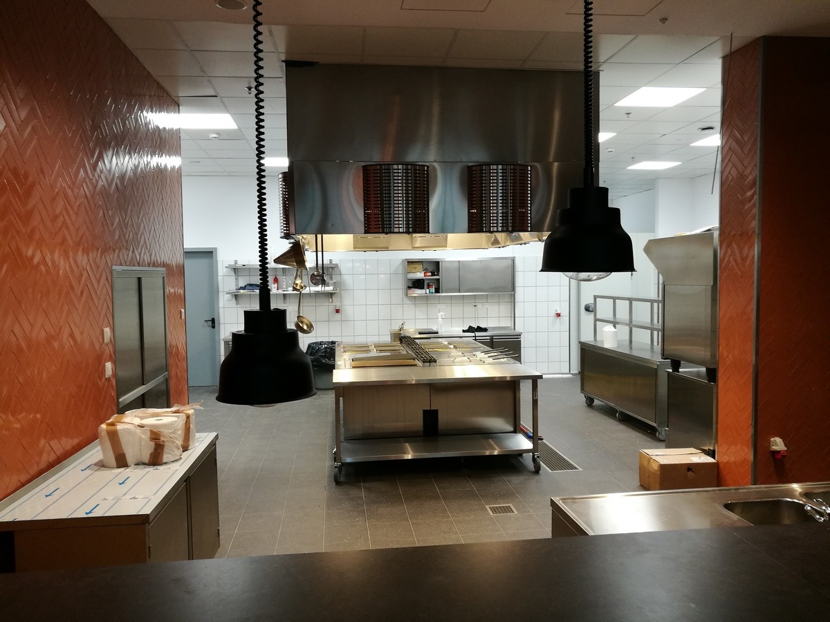 Profesjonalna kuchnia gastronomiczna ze stali nierdzewnej, wyposażona w centralny stół roboczy, okap wentylacyjny i wiszące lampy. Pomarańczowa ściana z wzorem w jodełkę po lewej stronie, białe...